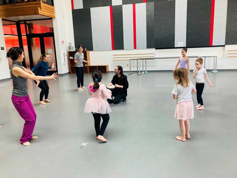 A group of dancing teachers and students stand in a circle. In the center of the circle, a musician sits on the floor and plays a hand drum.