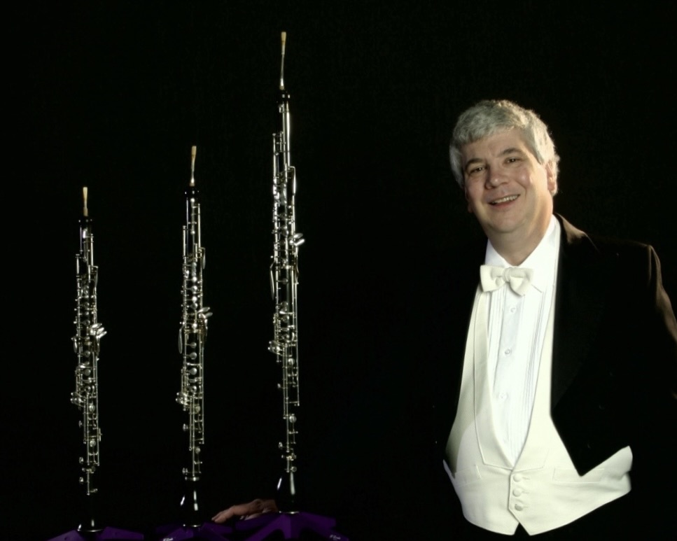 Tad Margelli, wearing a full tuxedo, stands smiling next to several wind instruments