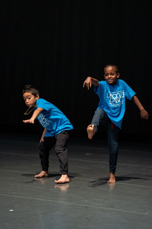 Two young boys of color dance energeticaly onstage. They are wearing bright blue T-shirts. One kicks his leg twoards the camera, while the other bends low to the ground and reaches forward.