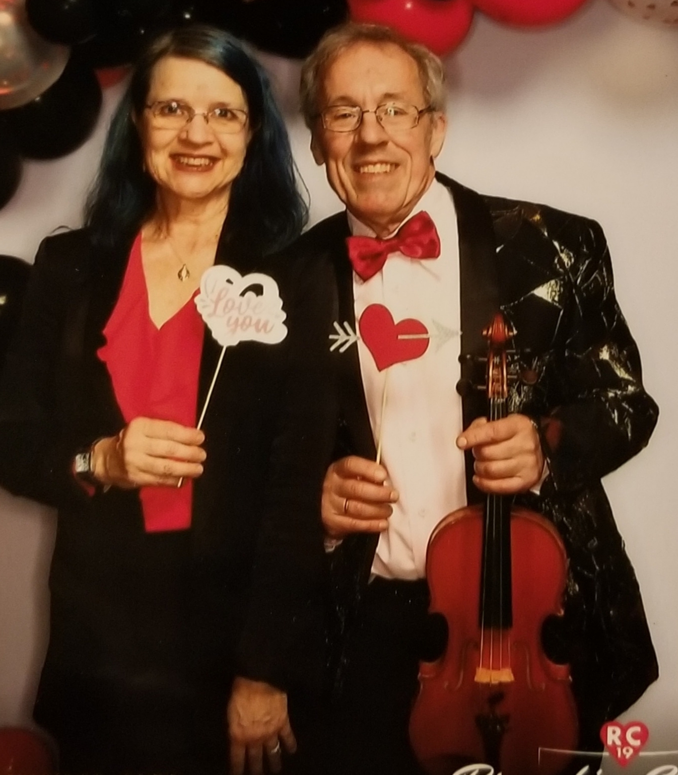 A smiling man wearing a red bowtie and a tux holds a violin. He is standing next to a woman in a red shirt and blazer, who is holding a small sign that reads, "I Love You."