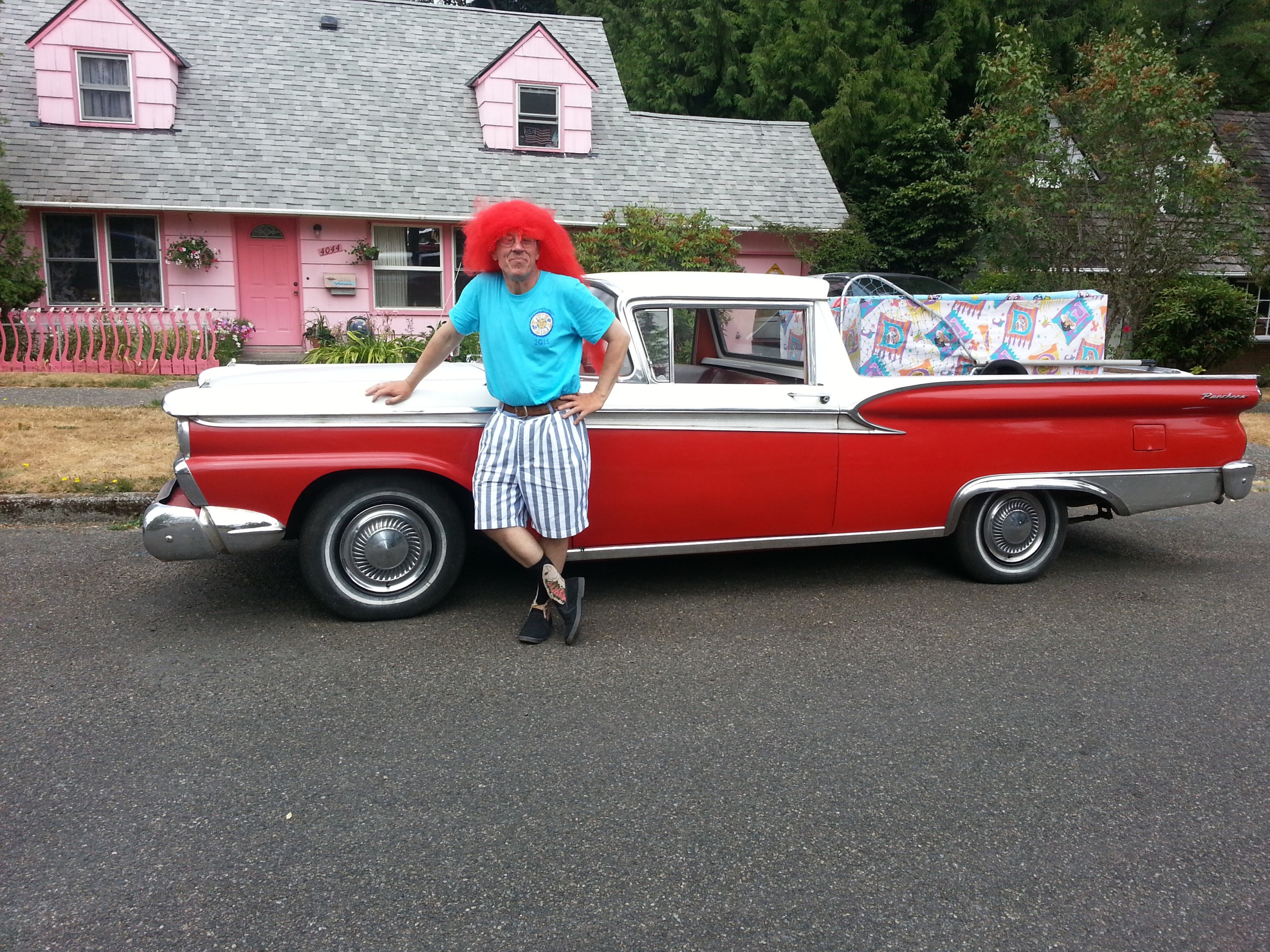 A man wearing an outrageous red wig leans agains a '59 Ford Ranchero.