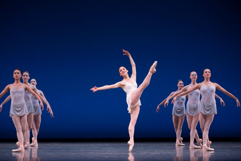 PNB Company dancer Leta Biasucci, wearing a white leotard, smiles and lifts her leg high across her body. Groups of other dancers wearing light blue leotards stand on either side of her, framing her as she dances.