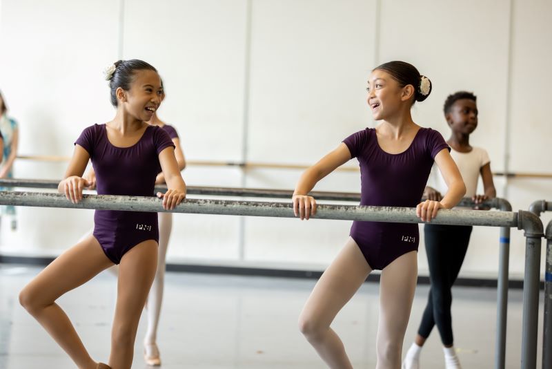 Two young girls standing at a ballet barre grin at each other.