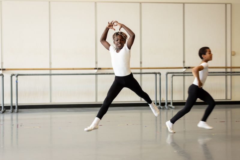 A young black boy leaps across a dance studio.