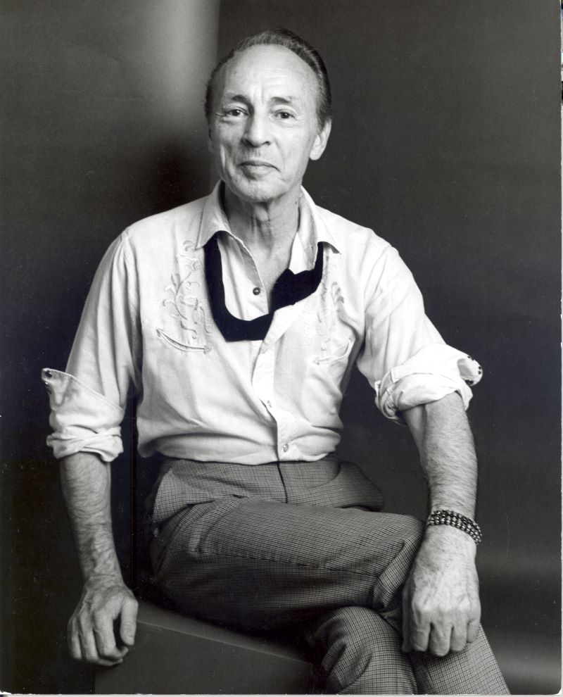 In this black and white portriat, ballet choreographer George Balanchine sits cross legged and looks directly at the camera. He is wearing a white button down shirt, trousers, and smiling softly.