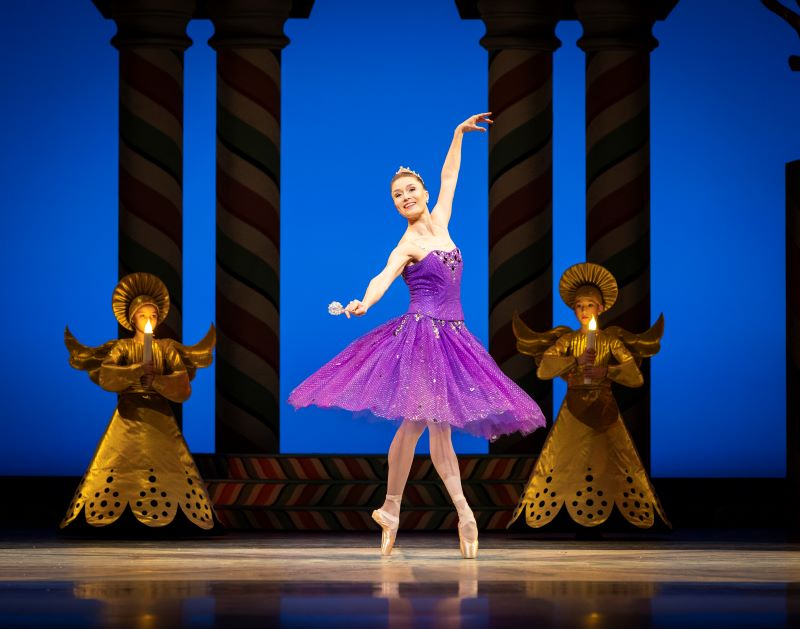 A dancer wearing a sparkly, flowing purple skirt and bodice appears to have just finished turning. Her skirt swirls out, and she reaches her arms gracefully outward. She is wearing a tiara and holding a wand. Behind her, two younger dancers wearing angel costumes and holding candles stand on either side of her. A deep blue backdrop with four colums frame the scene.