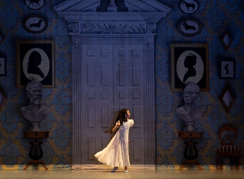 A young girl in a white nightgown stands onstage. The background behind her is dimly lit, and shows a large door with silhouette style portriats framing either side. The dancer is looking to the right of the stage, and appears to be just about to run that direction.