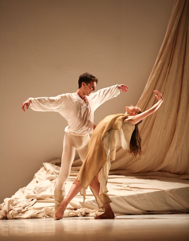 Two dancers perform a duet on stage against a backdrop of draped fabric. The male dancer, wearing a loose white shirt and white tights, holds his arms out as he gazes at his partner. The female dancer, in a beige, flowing dress, leans back gracefully with her arms extended, arching her body toward him. The soft lighting and neutral tones create an intimate and serene atmosphere.