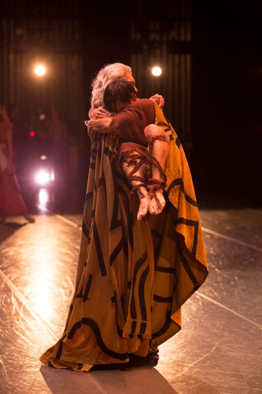 A dancer with long white hair wearing flowing yellow robes holds another dancer in their arms. The dancer being held wears tattered brown garments, and embraces the older dancer as they are held.