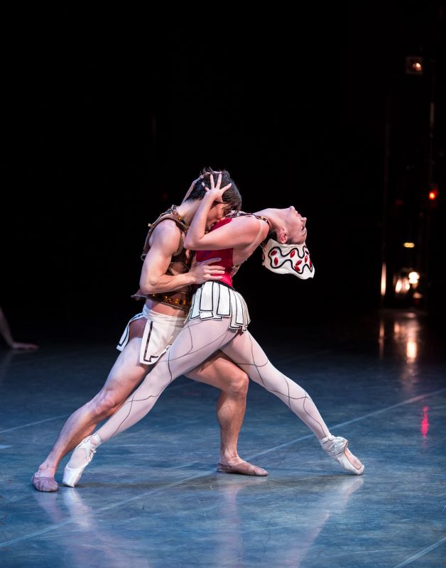 A male dancer wearing a white, pleated skirt stands in a deep lunge. He is embracing a female dancer wearing a red leotard, pleated skirt, white tights, and a dramatic headress. The male dancer burys his head into the female dancer's chest, and her head is thrown back.