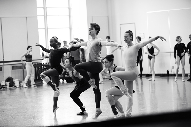 In this black and white photo, a group of dancers rehearse in a studio. Three of the dancers hold each others shoulders, each with one leg lifted twoards their chest and bent. Two other dancers duck under the groups arms. Behind them, other dancers observe them.