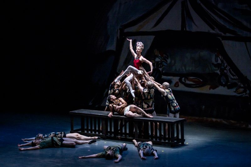 A group of dancers perform on a dimly lit stage. At the center, a dancer in a red top and white tights, wearing a large, ornate headdress, is held aloft by several others who are dressed in patterned costumes. They stand on or around a long, dark bench, with one dancer reclining across it. Other dancers lie on the floor around the bench with their arms outstretched, creating a dramatic tableau. A large, abstract backdrop looms behind them, adding to the theatrical atmosphere.