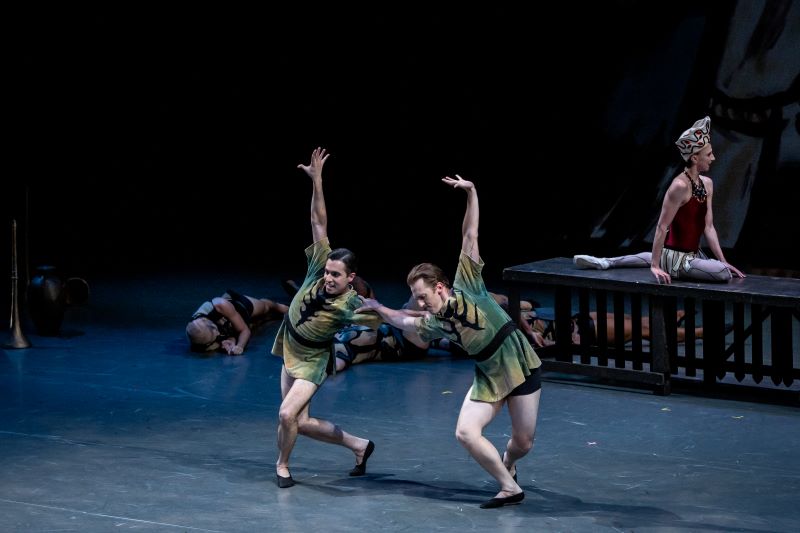 Two dancers wearing green tunics lean on each other. Their bodies are bent slightly forward as if moving in a playful manner, and they each have one arm extended over their heads. Behind them, a female dancer in a red leotard sits on a table, leaning twoards someone out of frame.