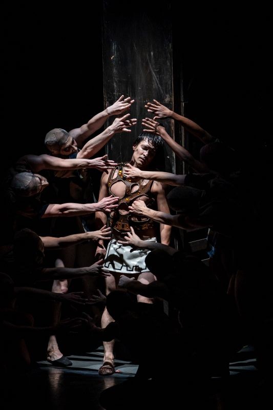 In this striking image, a dancer leans against a wooden post. Their eyes are closed, and their head hangs down exhausted. The stage around them is dimly lit, and many hands reach twoards them out of the darkness.