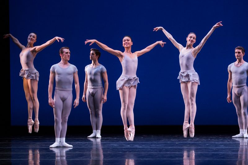 A group of dancers wear light colored leotards and tights. The stage background behind them is a deep blue. Three pointe dancers jump with their arms gracefully outstreched. Three other dancers in flat shoes stand behind them, observing.